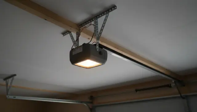 A belt-drive garage door opener hung from the ceiling joists, its light on